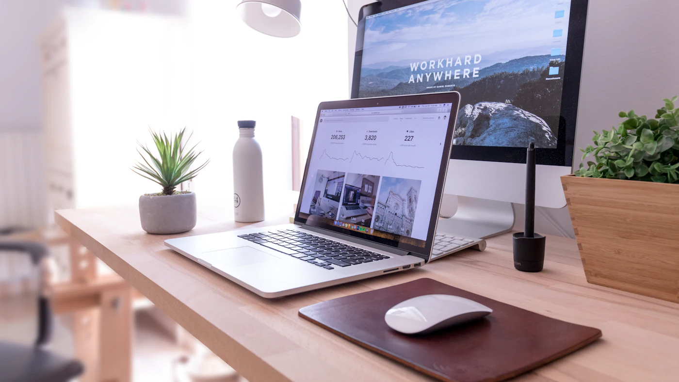 Meja kerja dengan monitor dan laptop yang menampilkan halaman website, lampu meja, dan tanaman