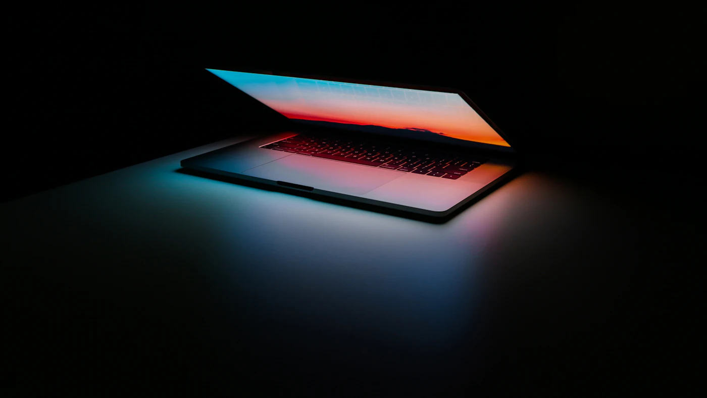 Laptop setengah tertutup memancarkan cahaya berwarna di ruangan gelap