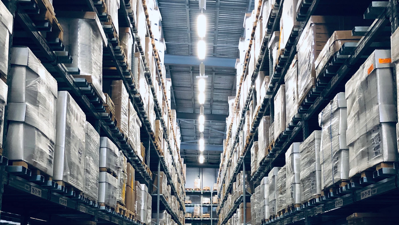 Lorong gudang dengan rak tinggi berisi tumpukan kardus